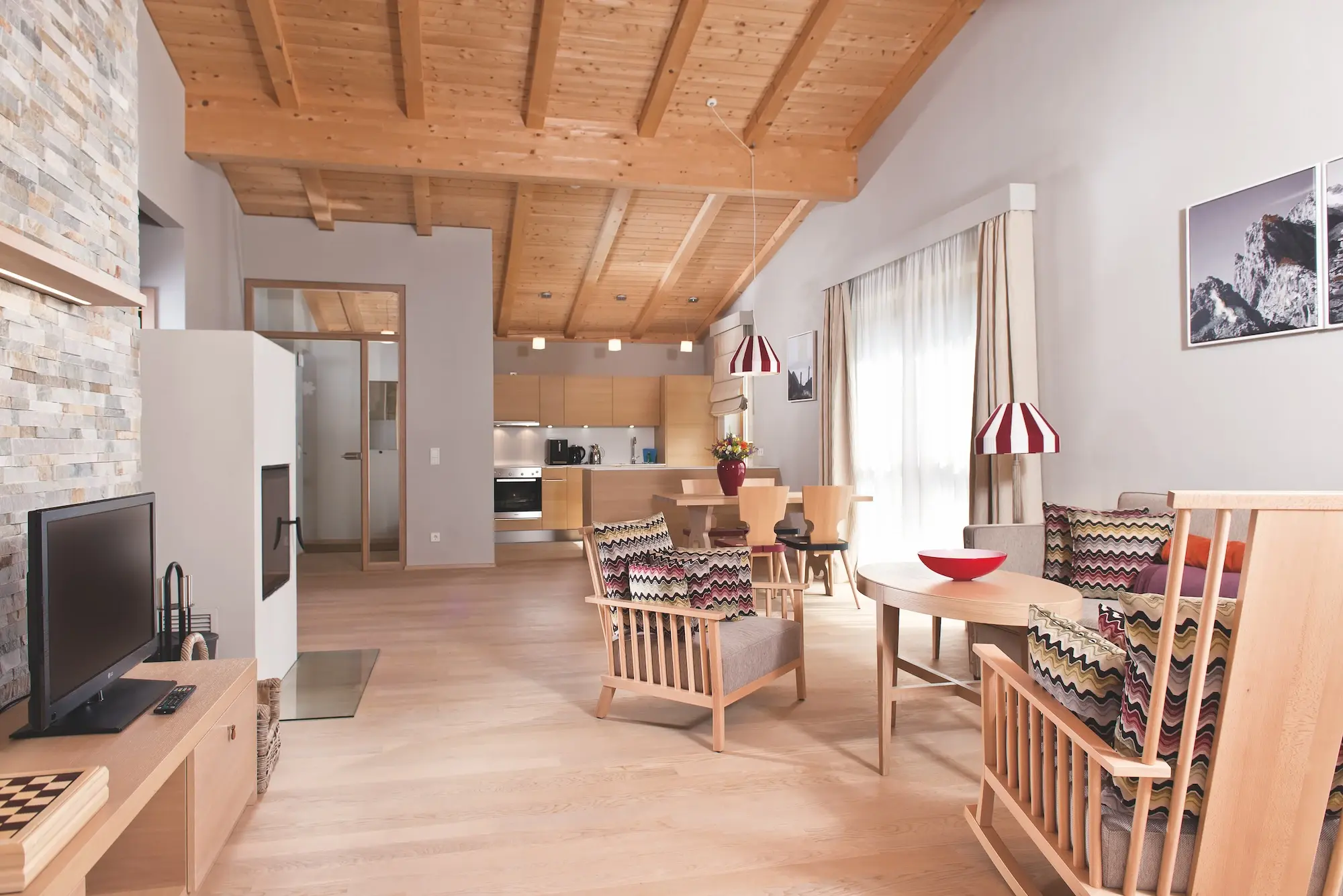 Interior of a vacation apartment with open kitchen and living room with wooden ceiling, table, chairs and a sofa.