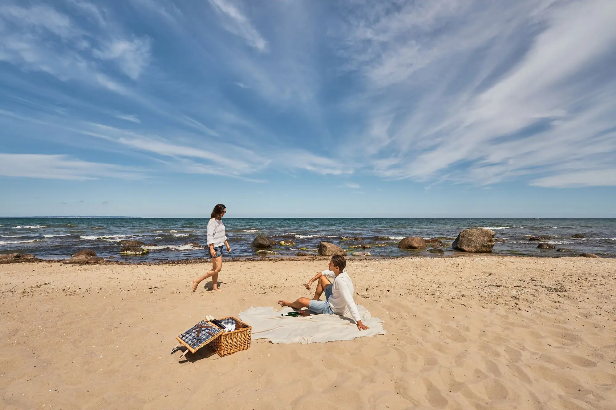 Göhren beach Couple having a picnic on a natural Baltic Sea beach; the man sits on a blanket in the sand, the woman walks barefoot along the water, in the foreground a picnic basket, in the background the sea with rocks and a distant horizon.