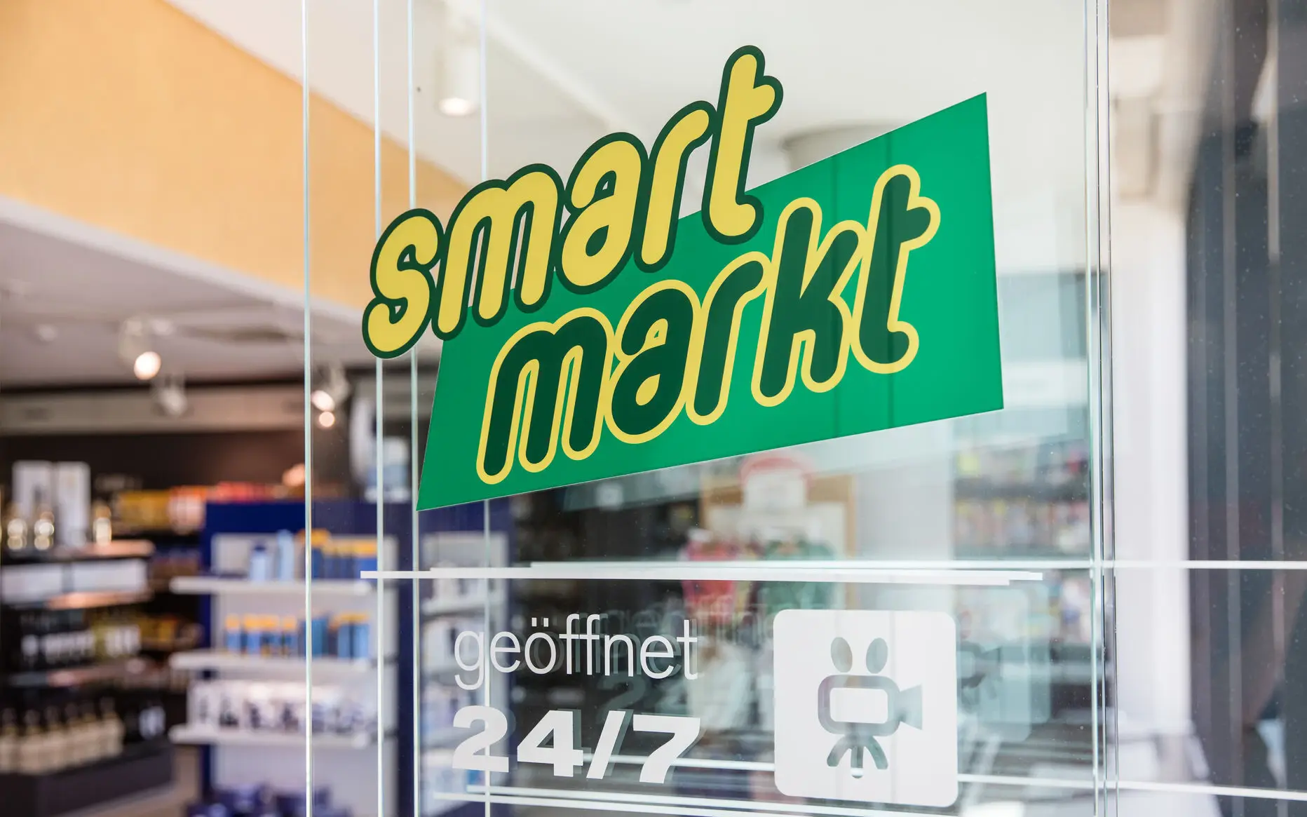 Glass door with the logo 'smart markt,' indicating a 24/7 open convenience store, with shelves of products visible in the background.