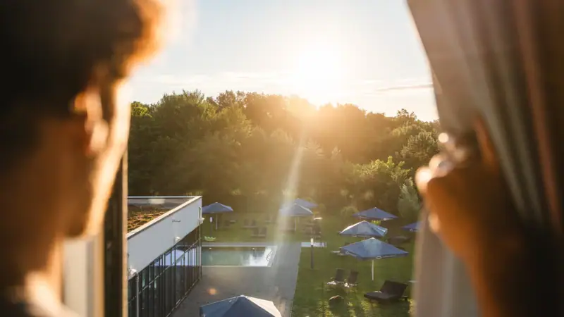 Rooms at the aja Bad Saarow A person looks at a pool with parasols and trees.