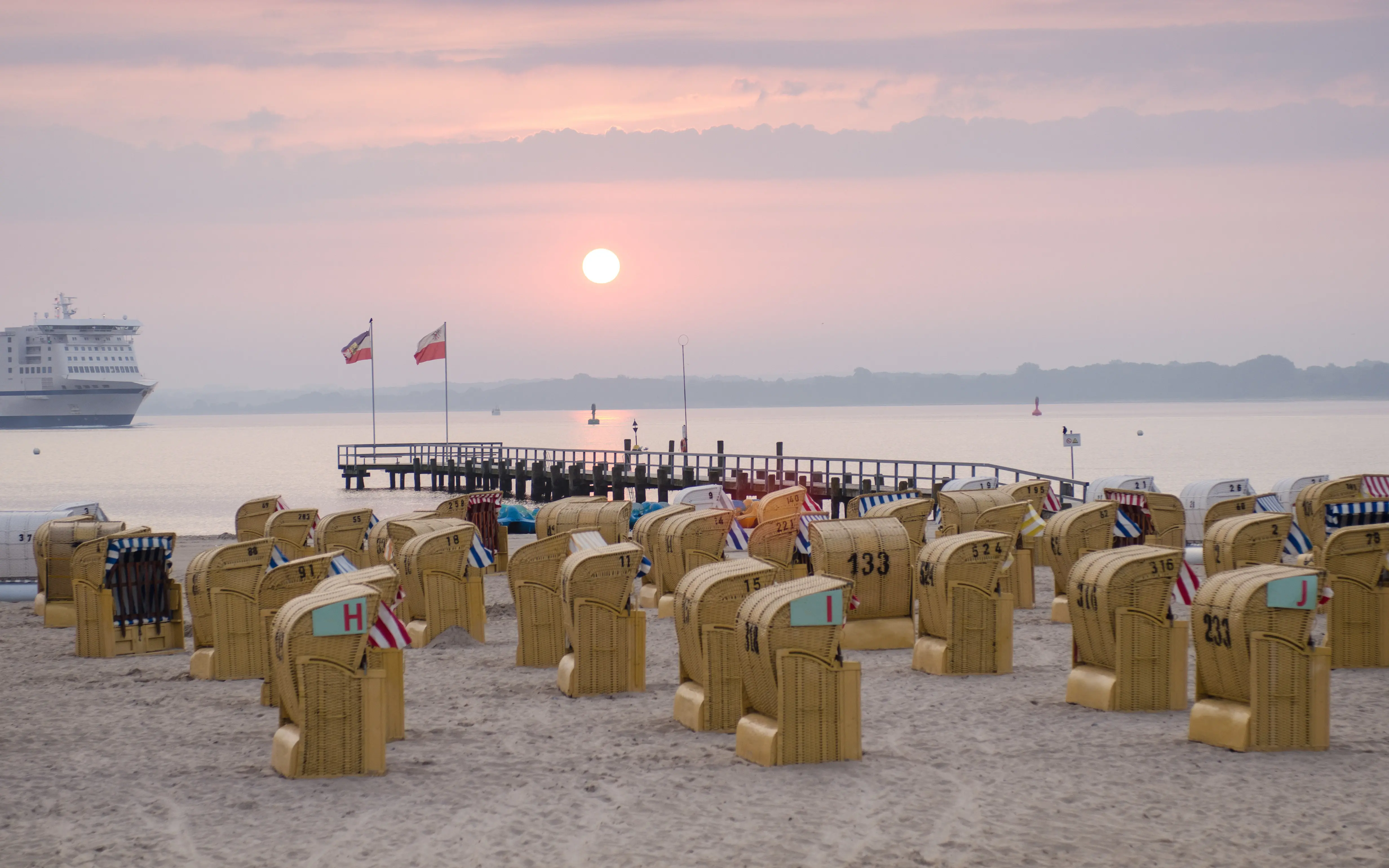 There are beach chairs on one section of the beach and a long jetty leads out onto the water. The sun sets in the sky