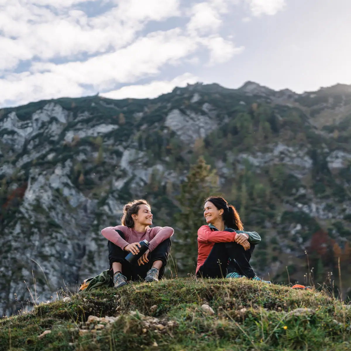 Hiking break Two women are sitting on a hill with mountains in the background.