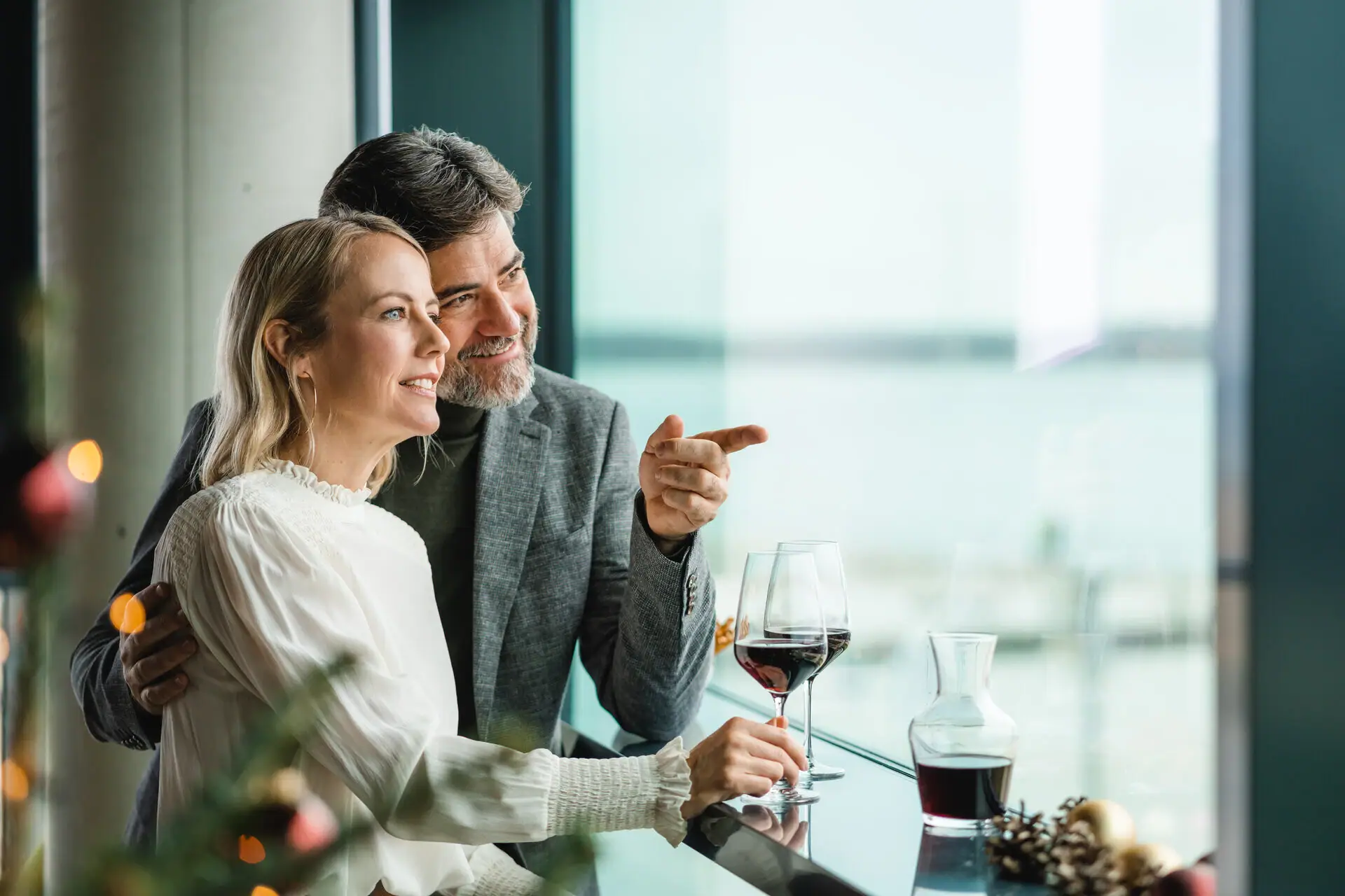A man and a woman sit at a bar with glasses of wine. Christmas lights shine in the foreground.