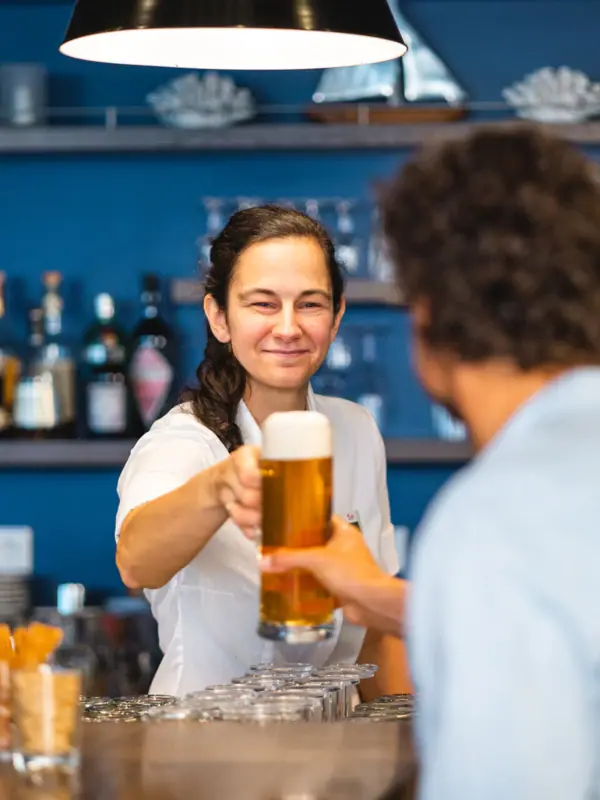 aja Bar Warnemünde Eine Frau hält ein Glas Bier an der Bar und übergibt es einem Gast in der aja Strandbar Warnemünde.