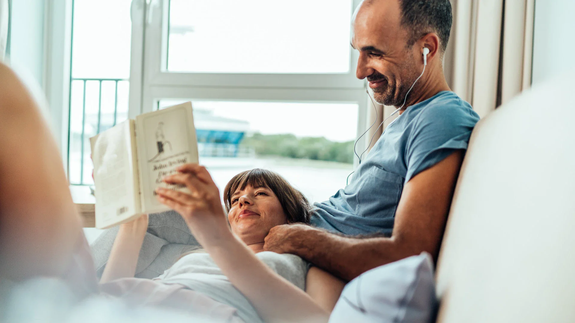 A cozy moment in a bright room with a couple relaxing on a couch; one person reading a book while the other listens to music with headphones, enjoying a peaceful atmosphere.