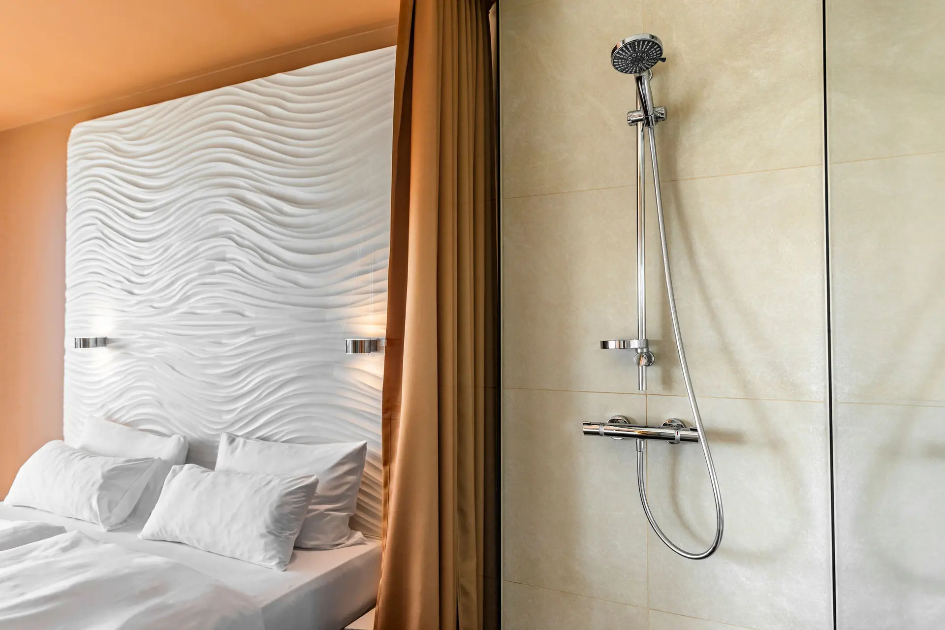 Stylish hotel room with a wave-patterned headboard and an open-plan design featuring a modern rainfall shower next to the bed, separated by elegant mustard-colored curtains.