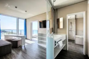 Suite at the aja Warnemünde Bathroom with shower and a glass wall, in the background the living area with TV and couch.