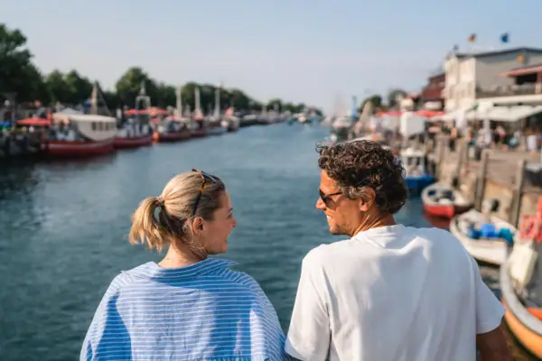 Alter Strom Warnemünde Ein Mann und eine Frau schauen sich an und im Hintergrund ist ein Fluss zu sehen..