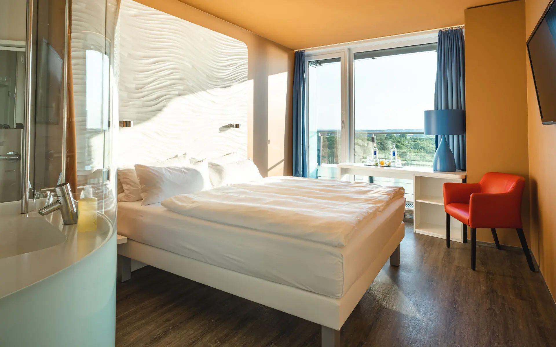 Bright and modern hotel room with a comfortable bed, wave-patterned headboard, large window offering a scenic view, and stylish decor including a red armchair and blue accents.