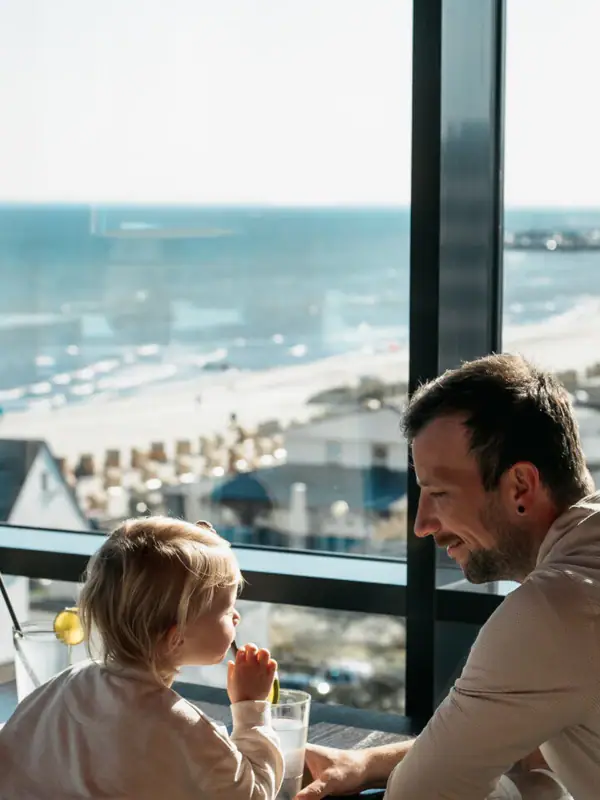 A man and a child are sitting at a table, looking out at a beach.