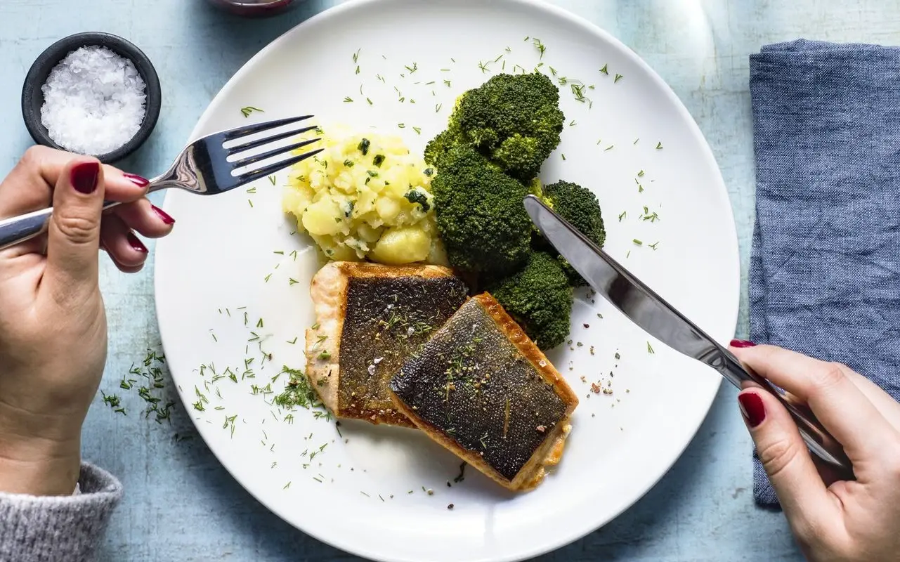 A person is sitting at a table with a fork in his hand, eating a plate of salmon, broccoli and mashed potatoes. 