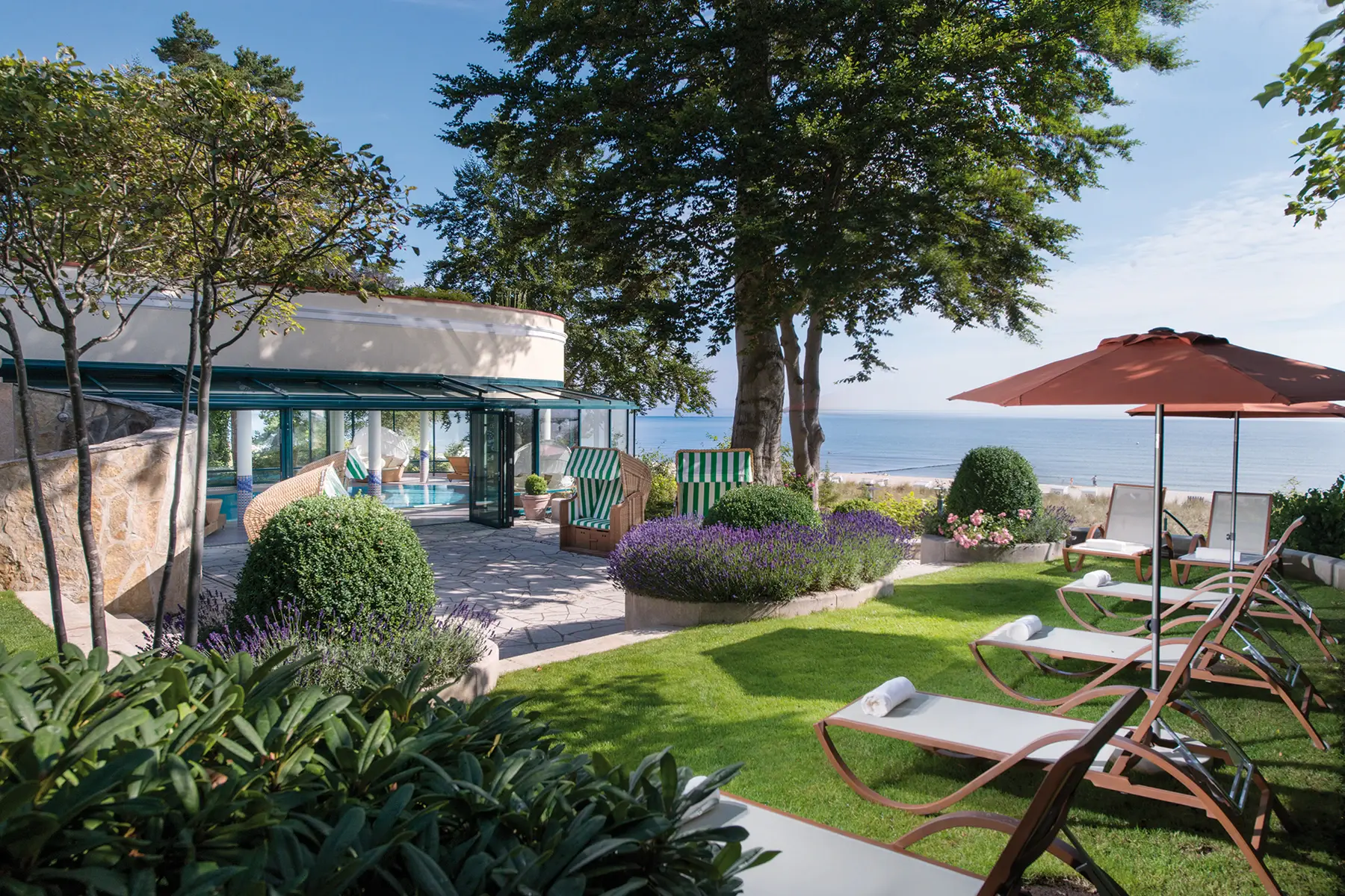 Deckchairs and a pool next to a building, surrounded by lush greenery and trees under a clear sky.