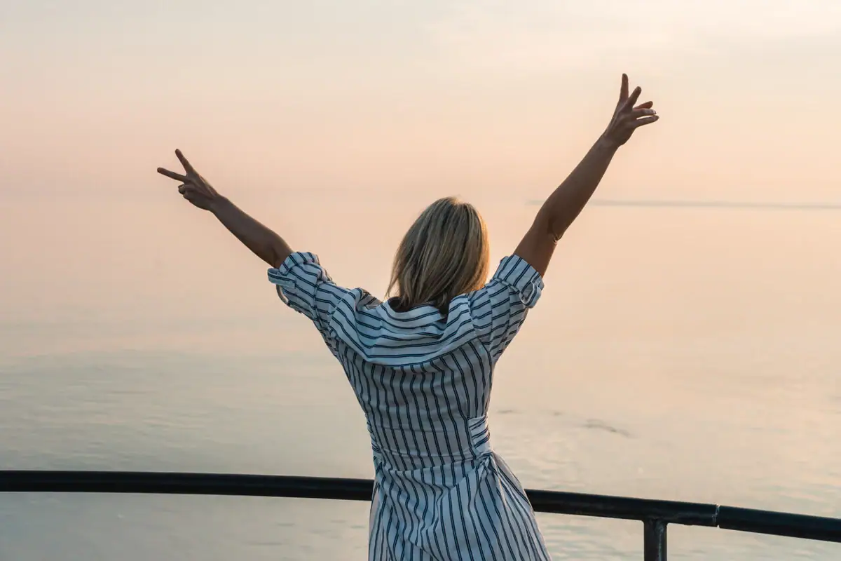 A woman stands on a railing with her arms raised and looks towards the sea.