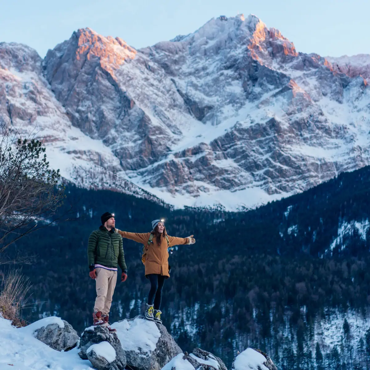 A man and a woman stand on a rock with snow and mountains in the background.