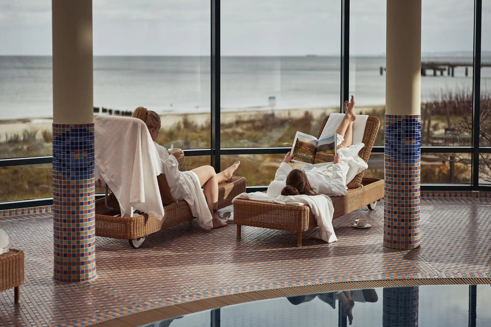 Women in bathrobes relax on sun loungers by a pool.