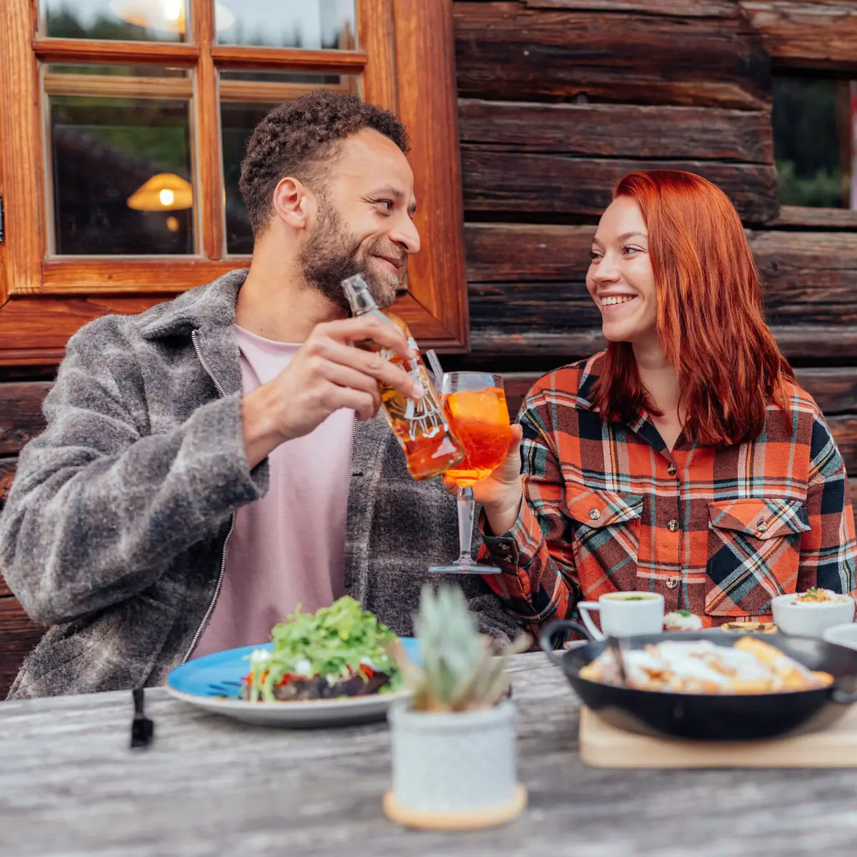 A man and a woman are drinking at a table with some food and a frying pan on it.