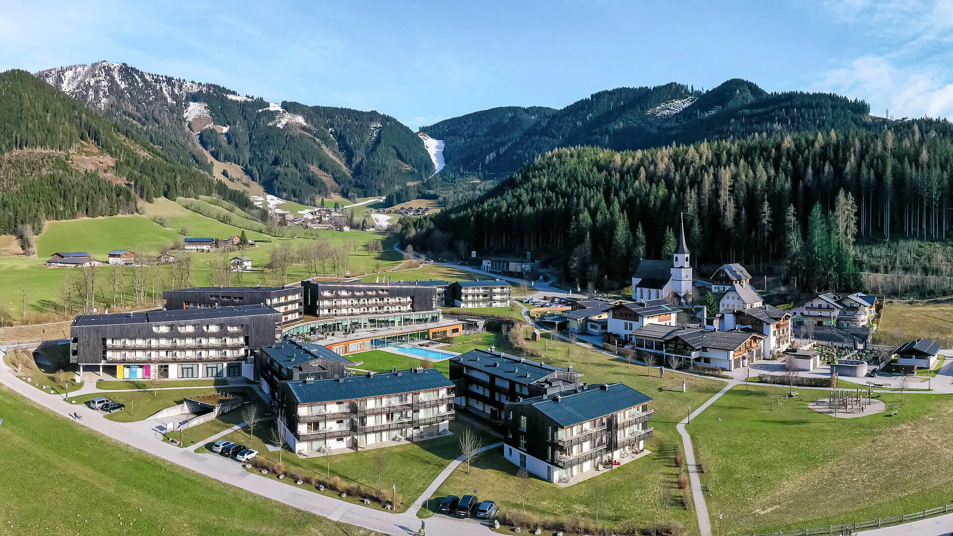A photograph of the aja Bergresort Werfenweng taken from above, with a church and mountains in the background.