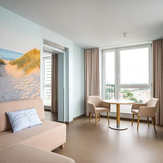 The living area of a hotel room, consisting of a light-coloured sofa and two chairs with a round table in front of a floor-to-ceiling window. Long beige curtains hang down from the window and a large picture of dunes, beach and sea hangs behind the sofa.