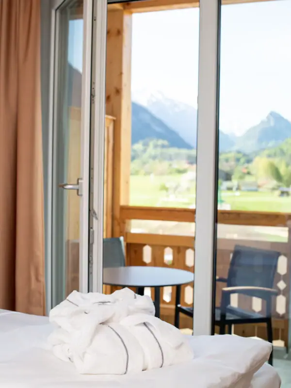 Room aja Ruhpolding A white bathrobe lies on a bed in front of an open balcony window with a view of the mountains.