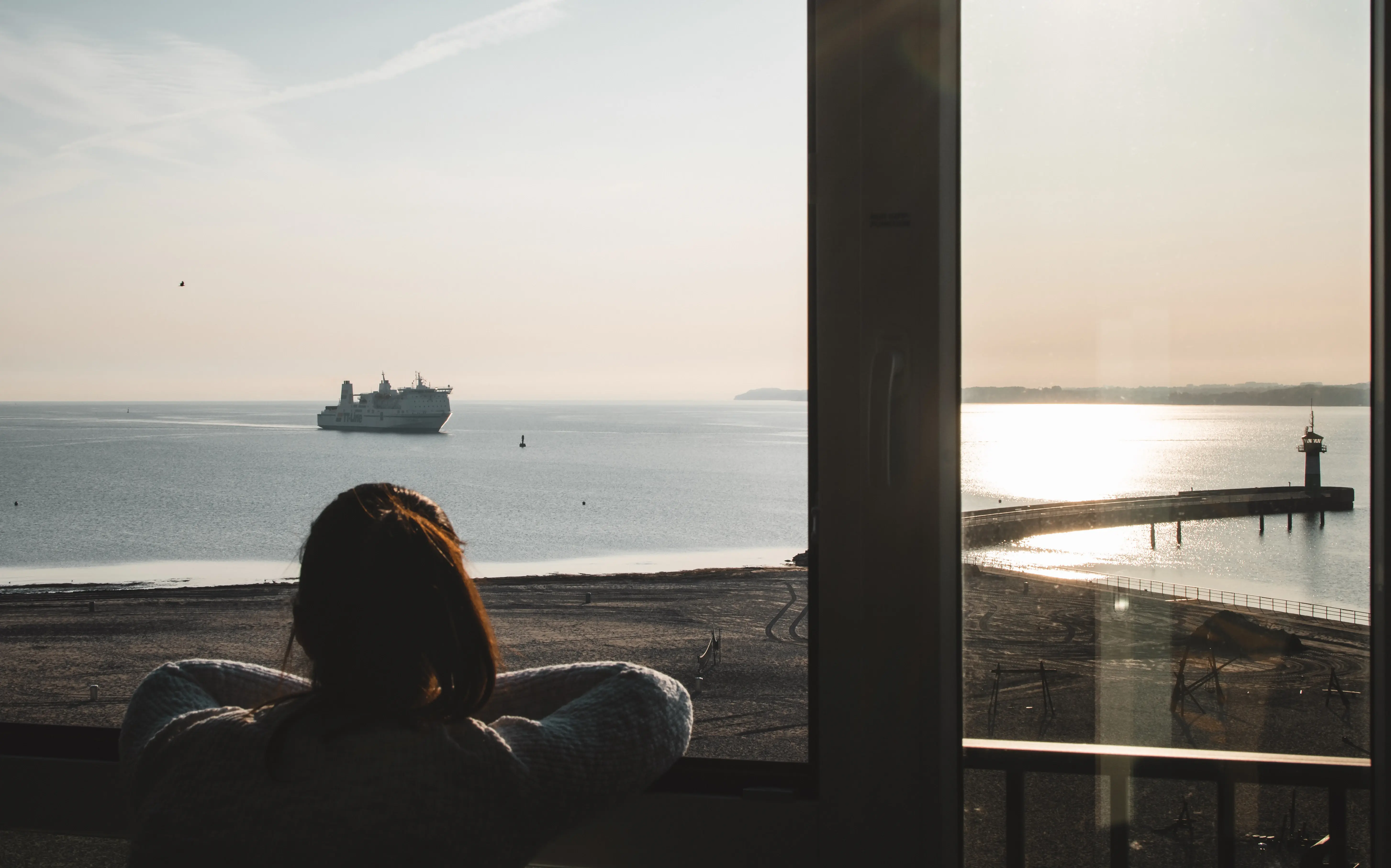 View from a window overlooking the sea, with a ship passing by and a lighthouse illuminated by sunlight.