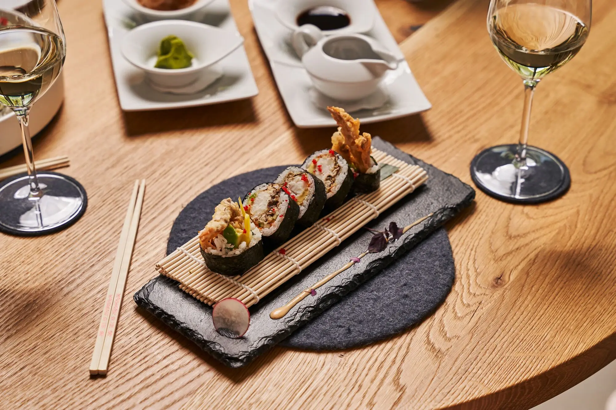 A plate of sushi on a table, surrounded by chopsticks and wine glasses.