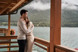 A man and a woman are embracing on a veranda overlooking a lake.
