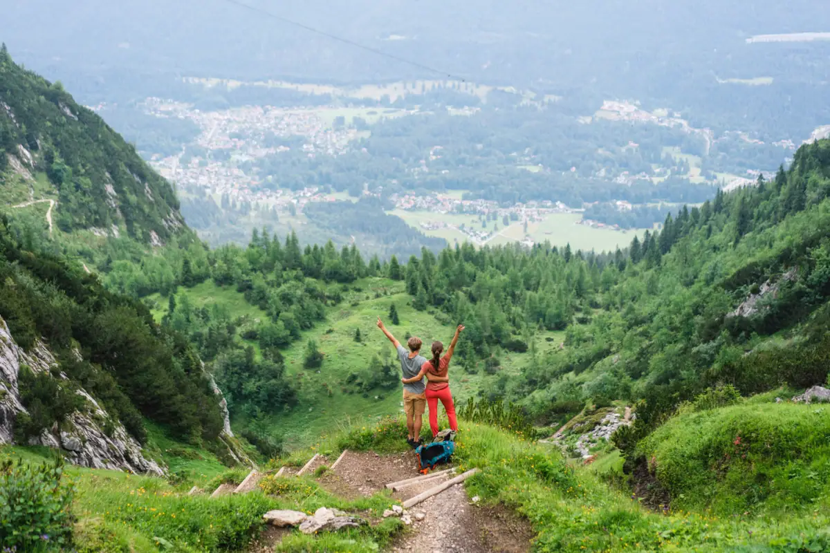 A man and a woman stand on a mountain during a hike and look down into the valley, stretching their arms in the air. 