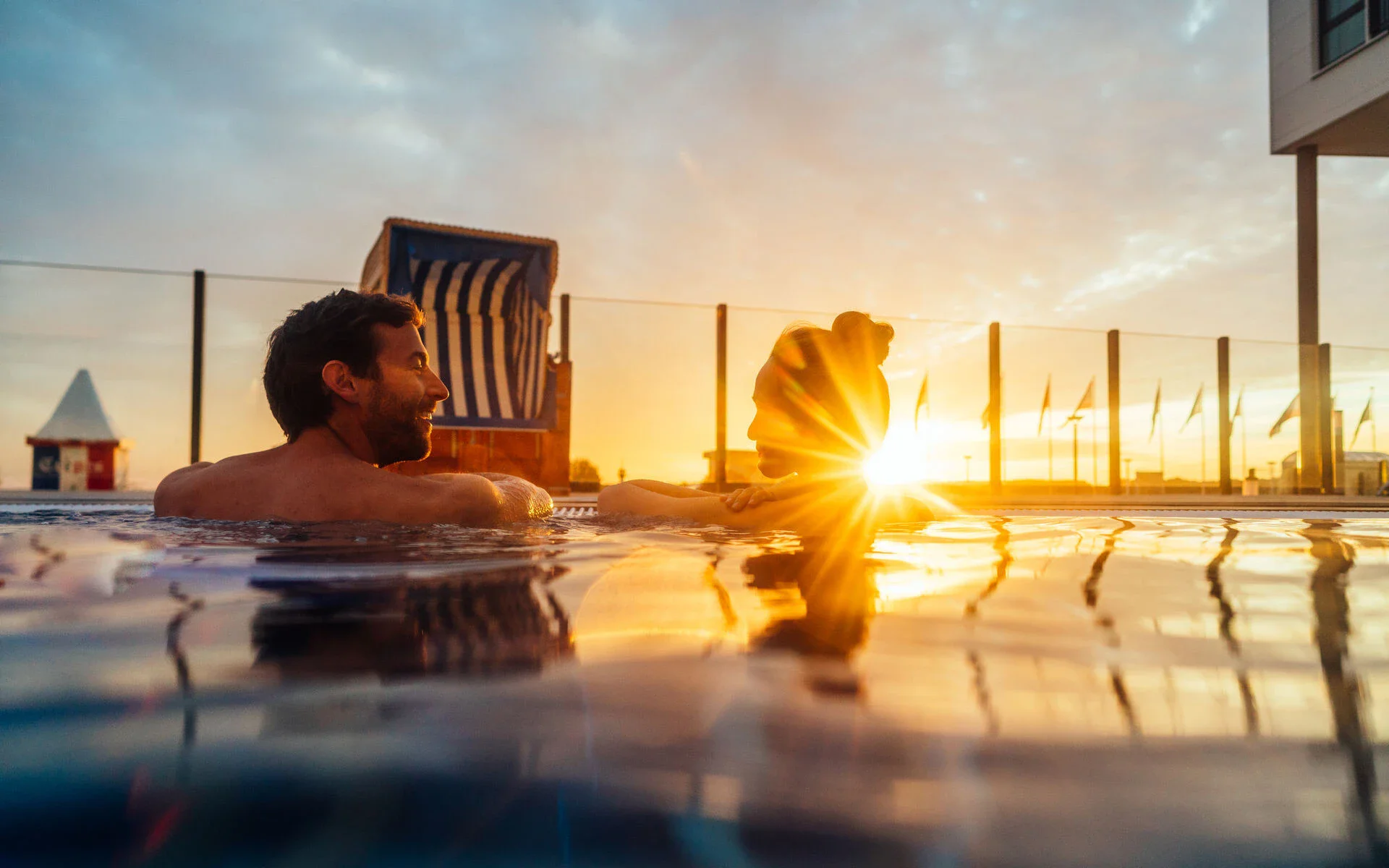 A couple relaxing in an outdoor pool at sunset, with warm sunlight reflecting on the water and a beach chair in the background, creating a serene and romantic atmosphere.