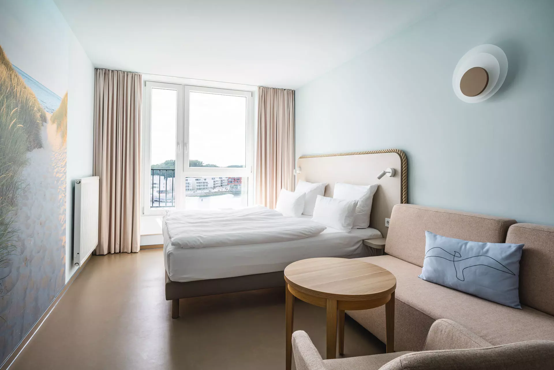 A bright room with a bed in front of a window and a couch in the foreground 