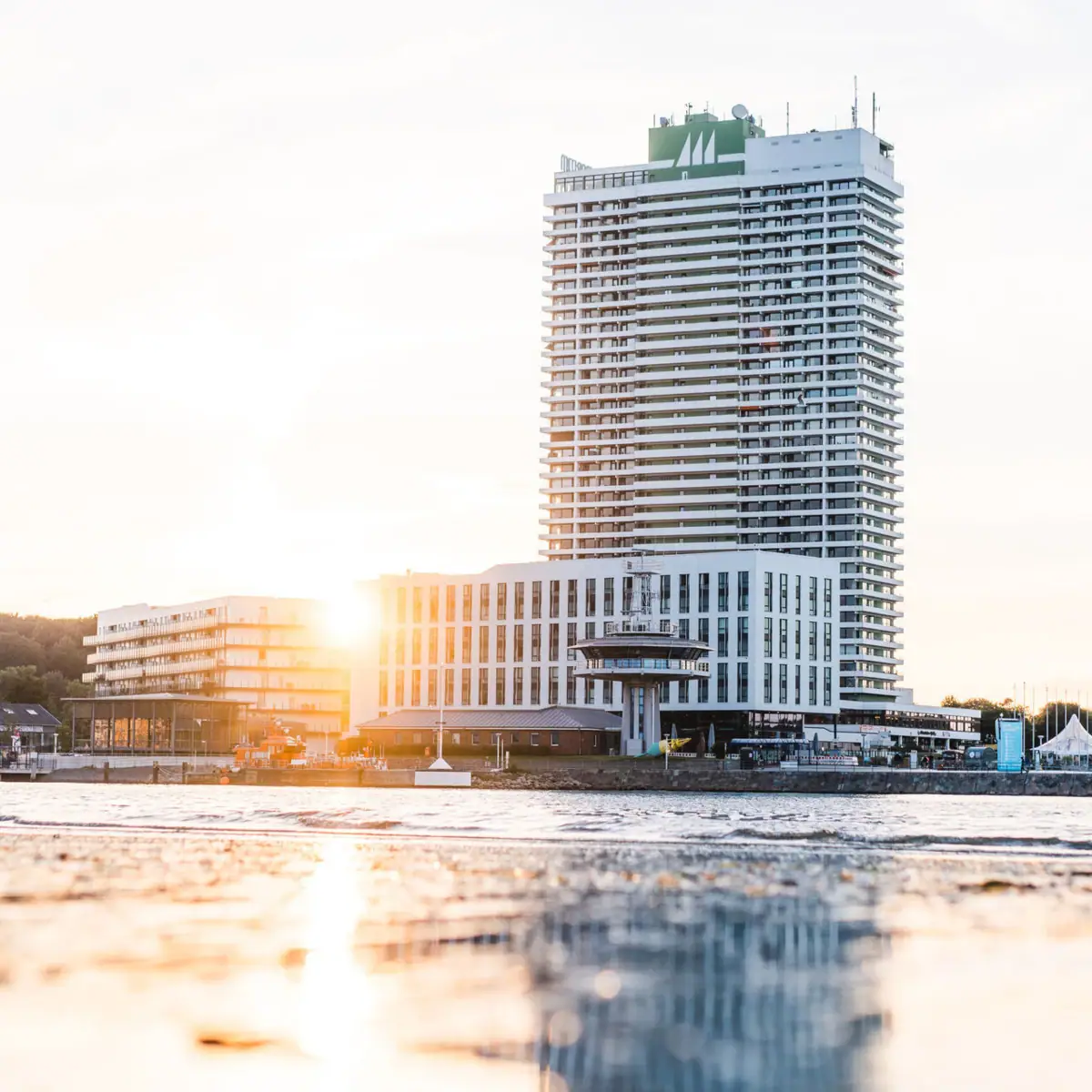 A large square hotel complex with many windows right by the sea. The morning sun shines powerfully from behind the hotel and is reflected in the shallow water.