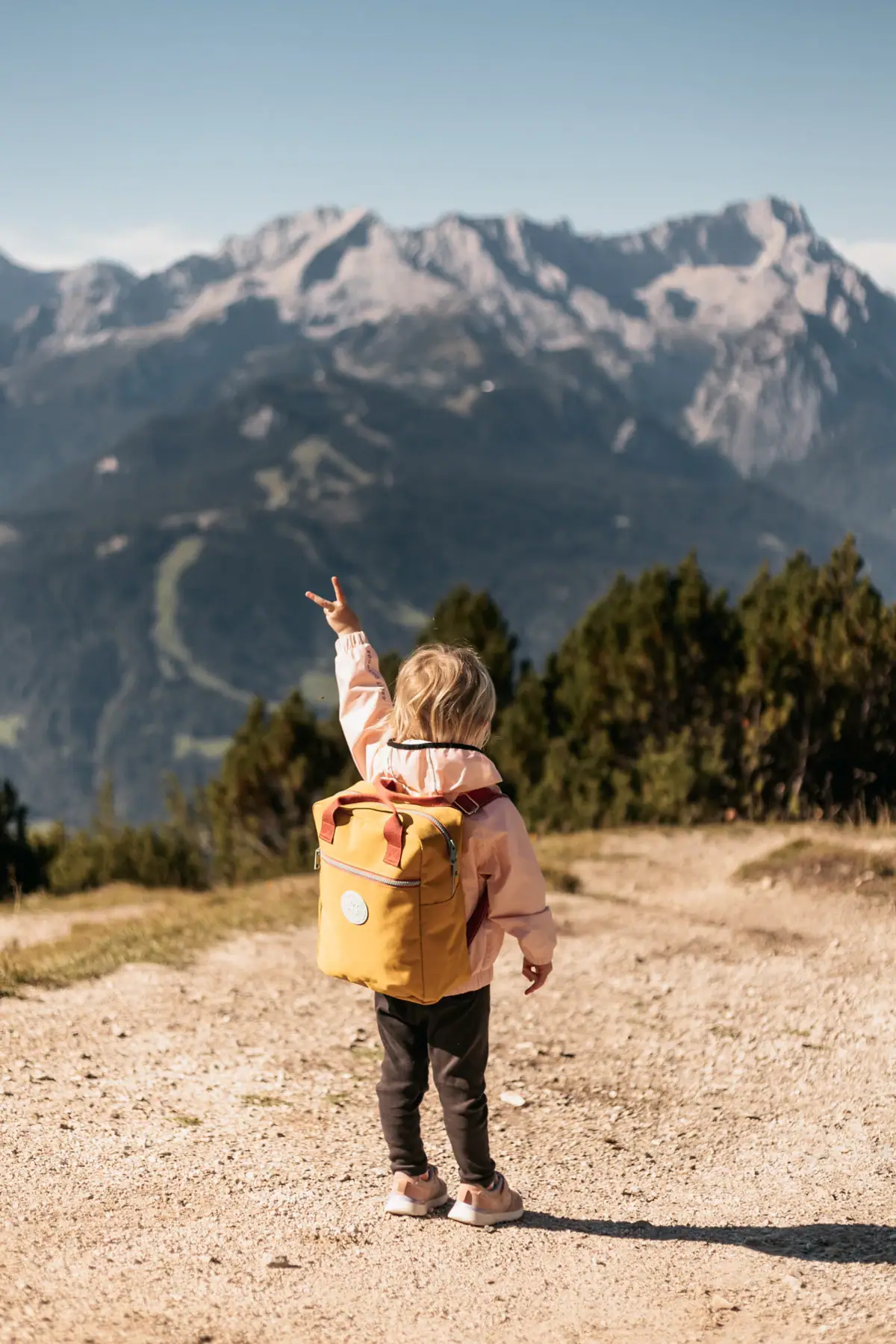 A child with a yellow rucksack stands on a dirt track in front of a mountain range.