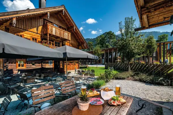 Cosy beer garden in front of a traditional alpine wooden hut with rustic charm in Werfenweng. Wooden tables and chairs under large parasols invite you to linger. A beer glass, a snack platter with sausage, cheese and fresh vegetables and a laid table are in the foreground.