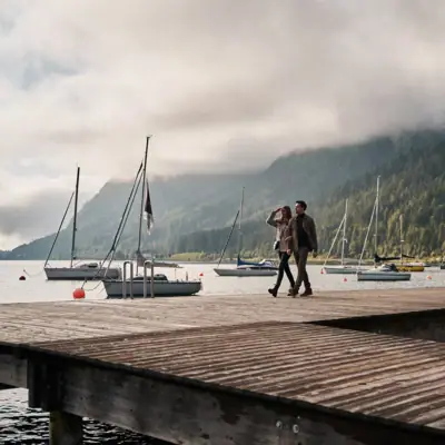 A couple walking on a dock with boats in the background.