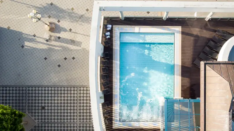 A pool on a terrace and a tiled area beneath it in the sun from a bird's eye view.