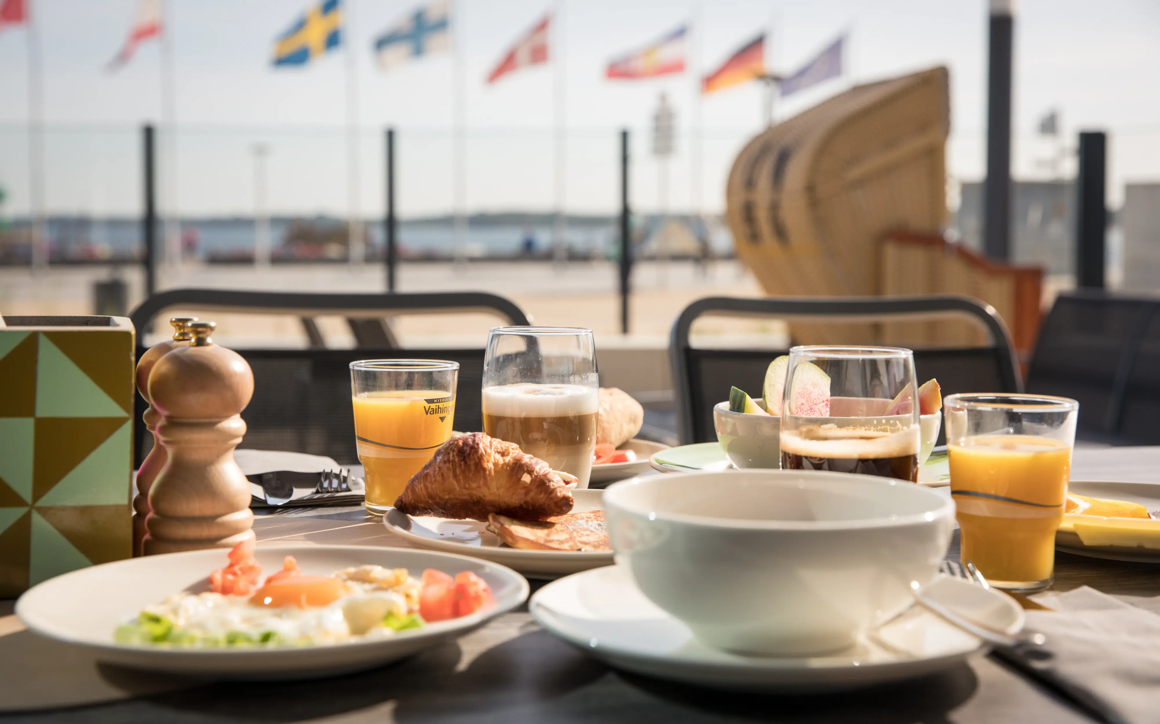 Delicious breakfast spread on an outdoor table, including a croissant, eggs, fresh fruit, and orange juice, with flags waving in the background and a beach chair in view.