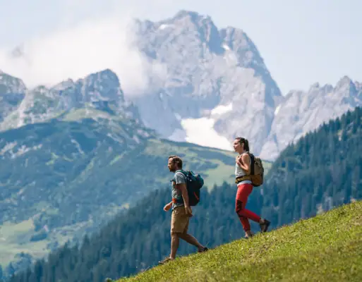 Two people walking on a grassy hill with mountains in the background.