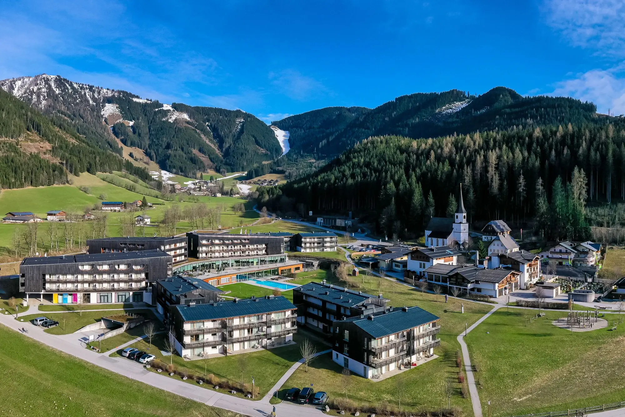 The Werfenweng mountain resort in a valley with mountains in the background, surrounded by grass, trees and clouds.