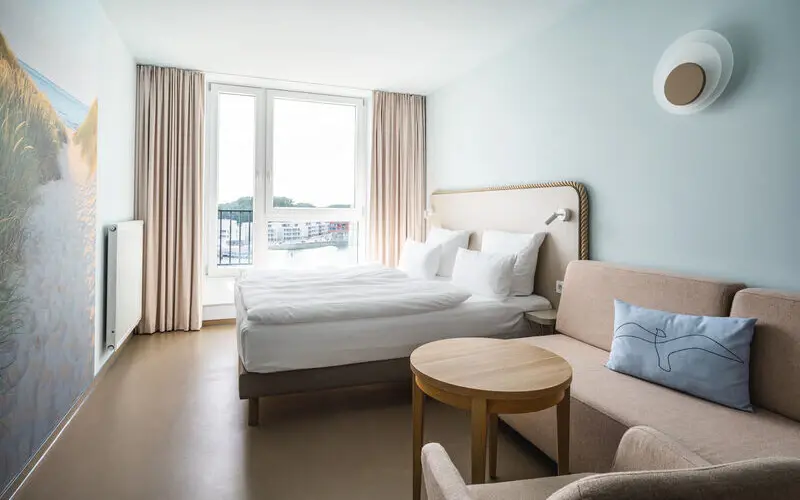 A bright hotel room featuring a double bed, beige sofa, light wooden furniture, and a large window with views of a nearby harbor.