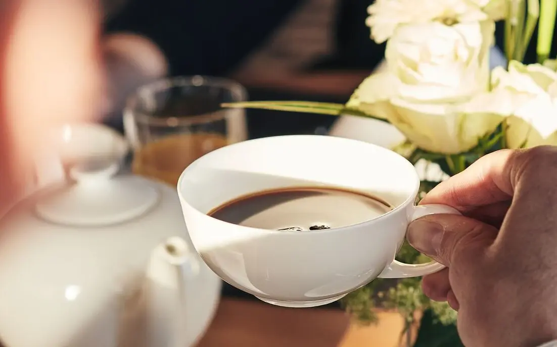 Person holding a cup of coffee with a pot and roses in the background.