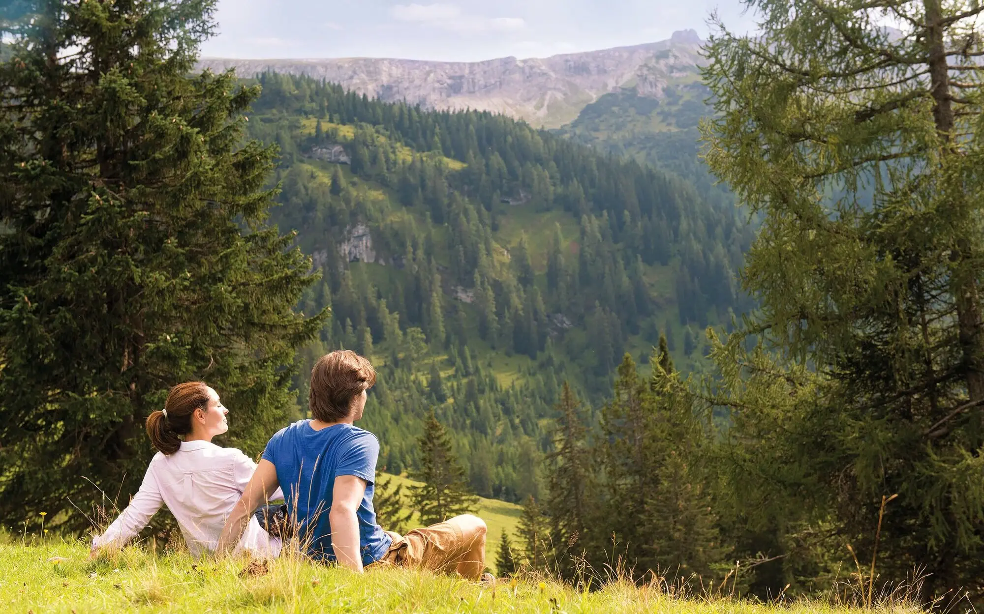 A man and a woman sit on a hill and look out over a mountain panorama.