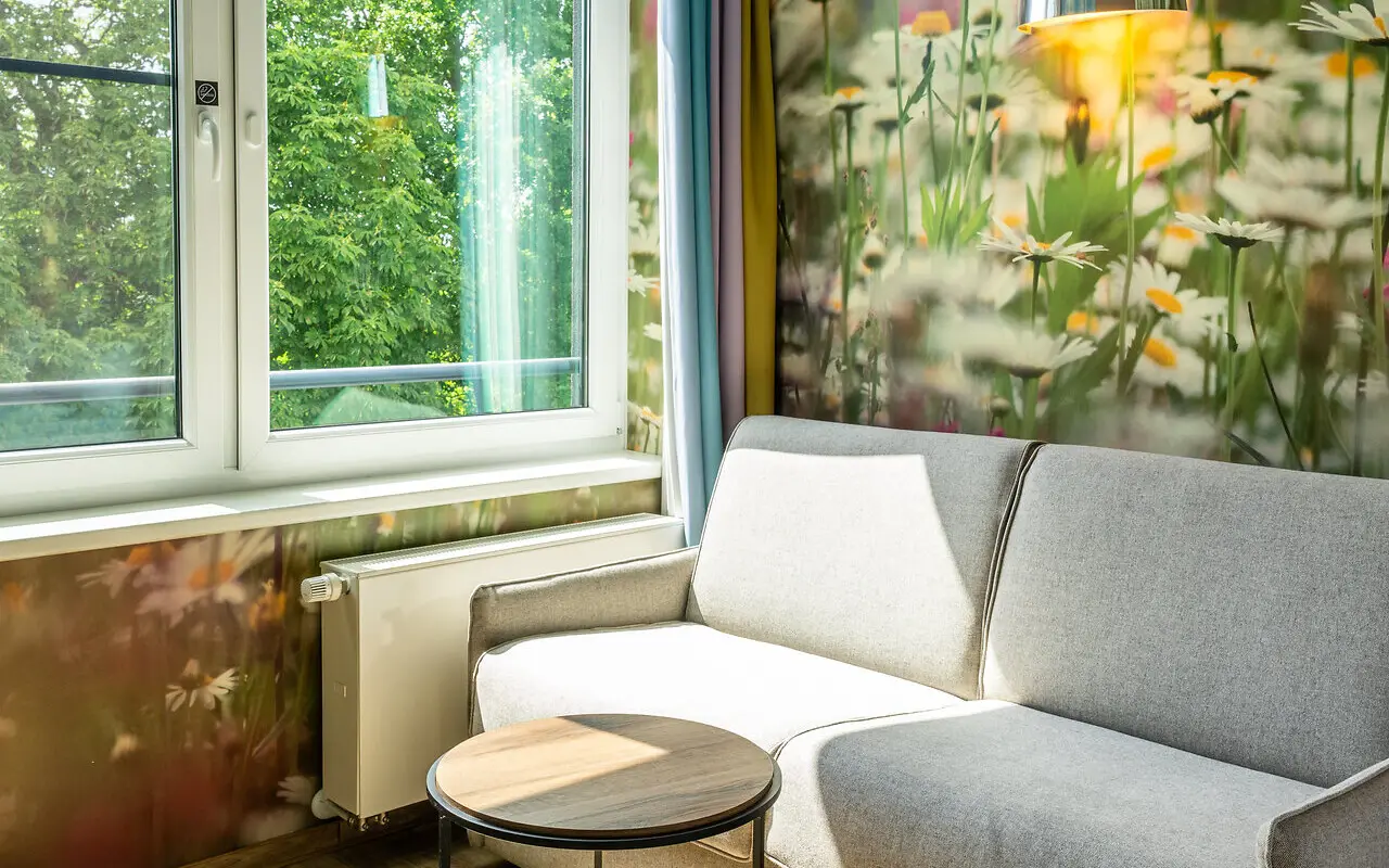 A cozy living space with a grey sofa and small round table, nature realistic flower wallpapers and a big window that fills the room with daylight. 