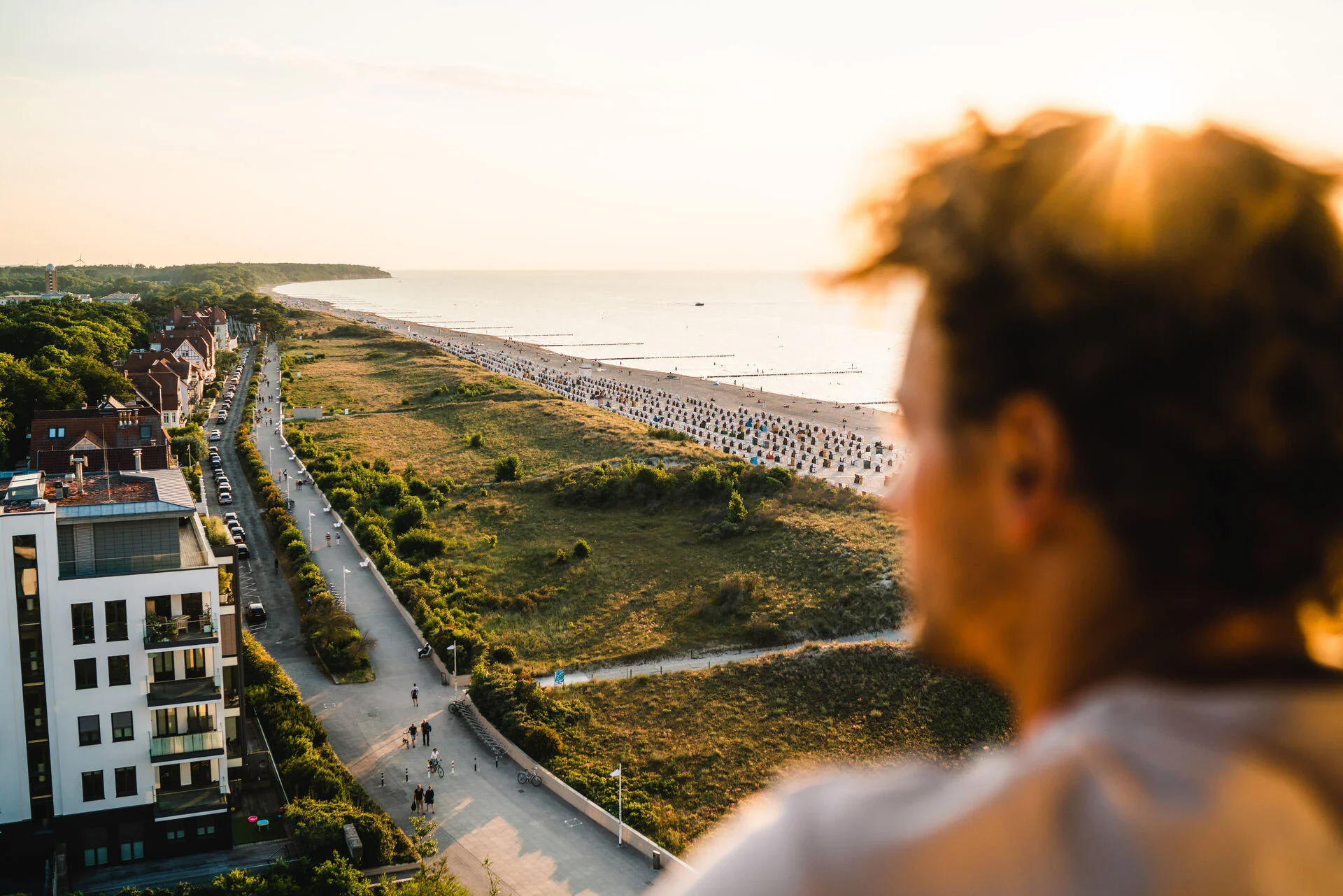 View from a balcony overlooking the beach promenade and dunes at sunset, with rows of beach chairs along the Baltic Sea and a person in the foreground enjoying the serene atmosphere.