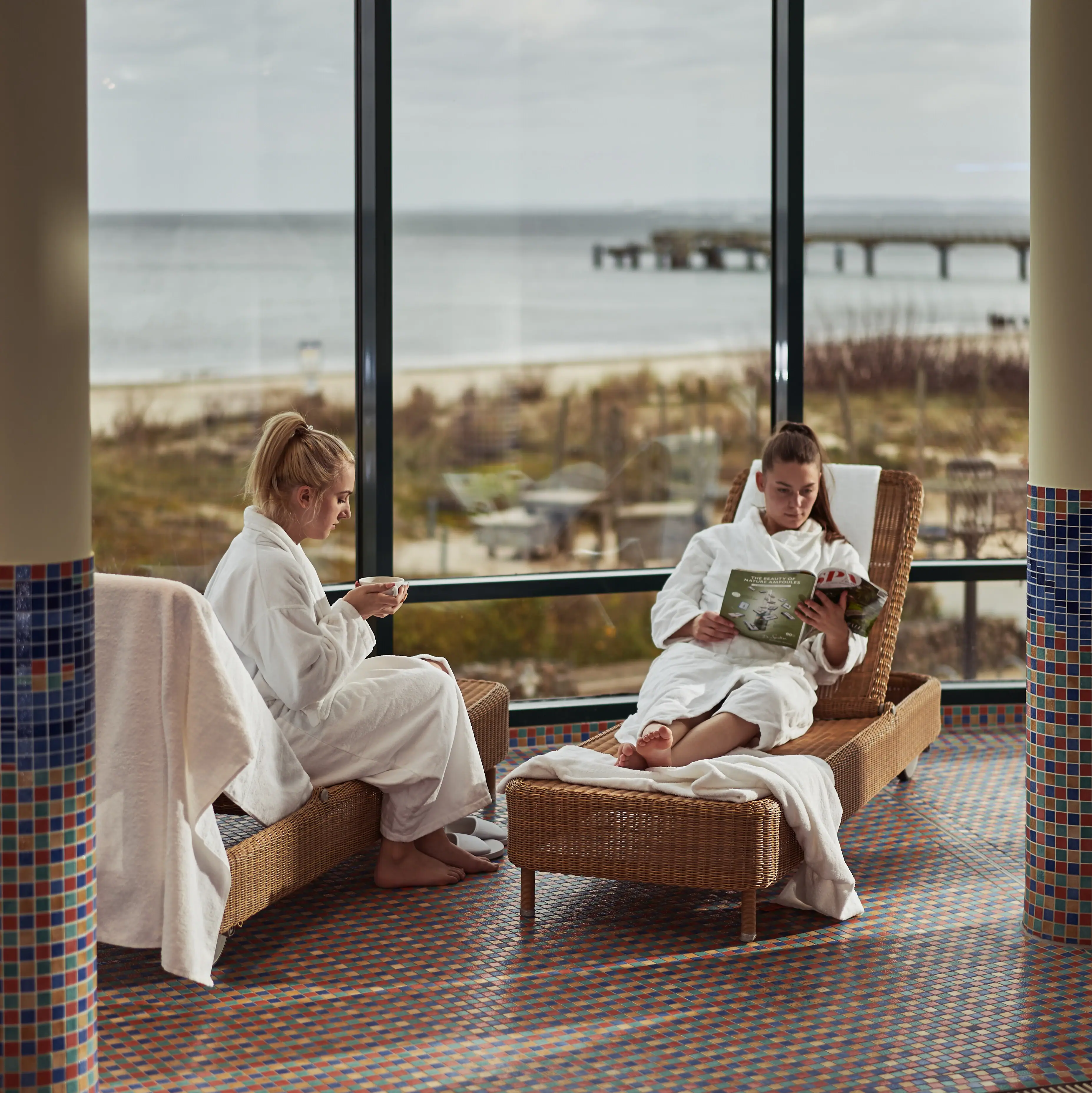 Two women in white bathrobes sit on chairs by the pool.