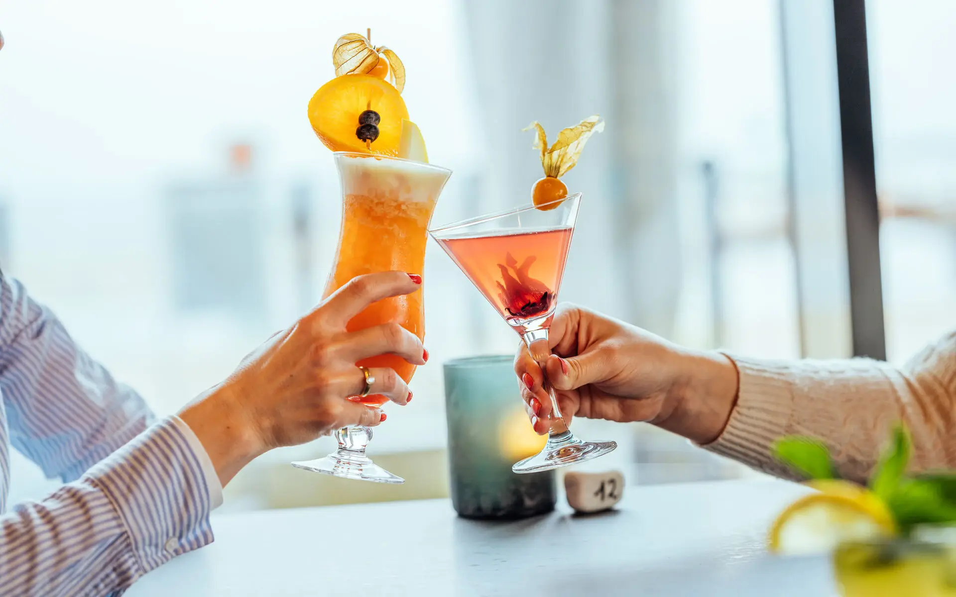 Two people sit at a bar counter and clink their cocktail glasses.