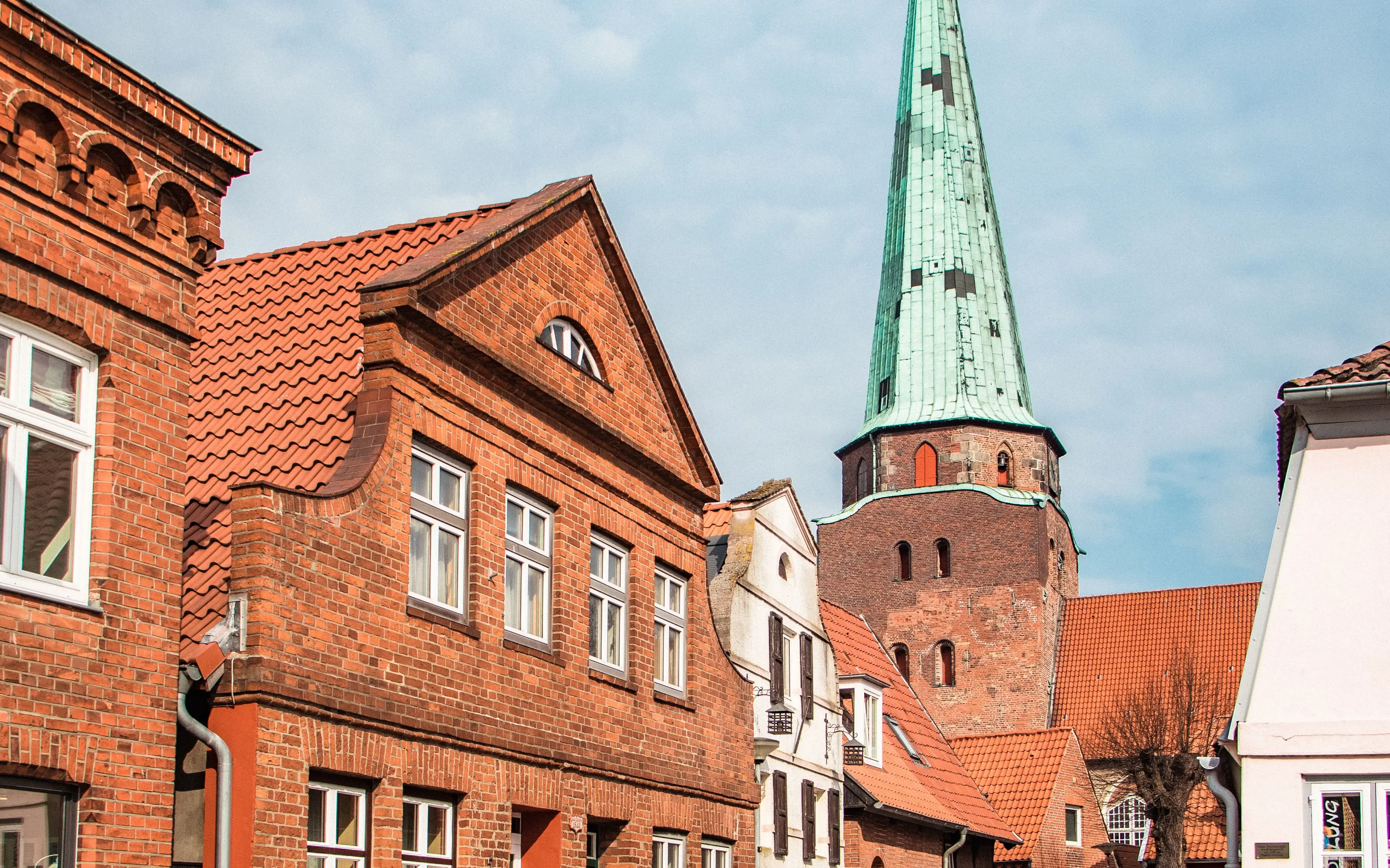 Charming cobblestone street lined with historic red-brick houses, leading to a church with a striking green spire under a partly cloudy sky.