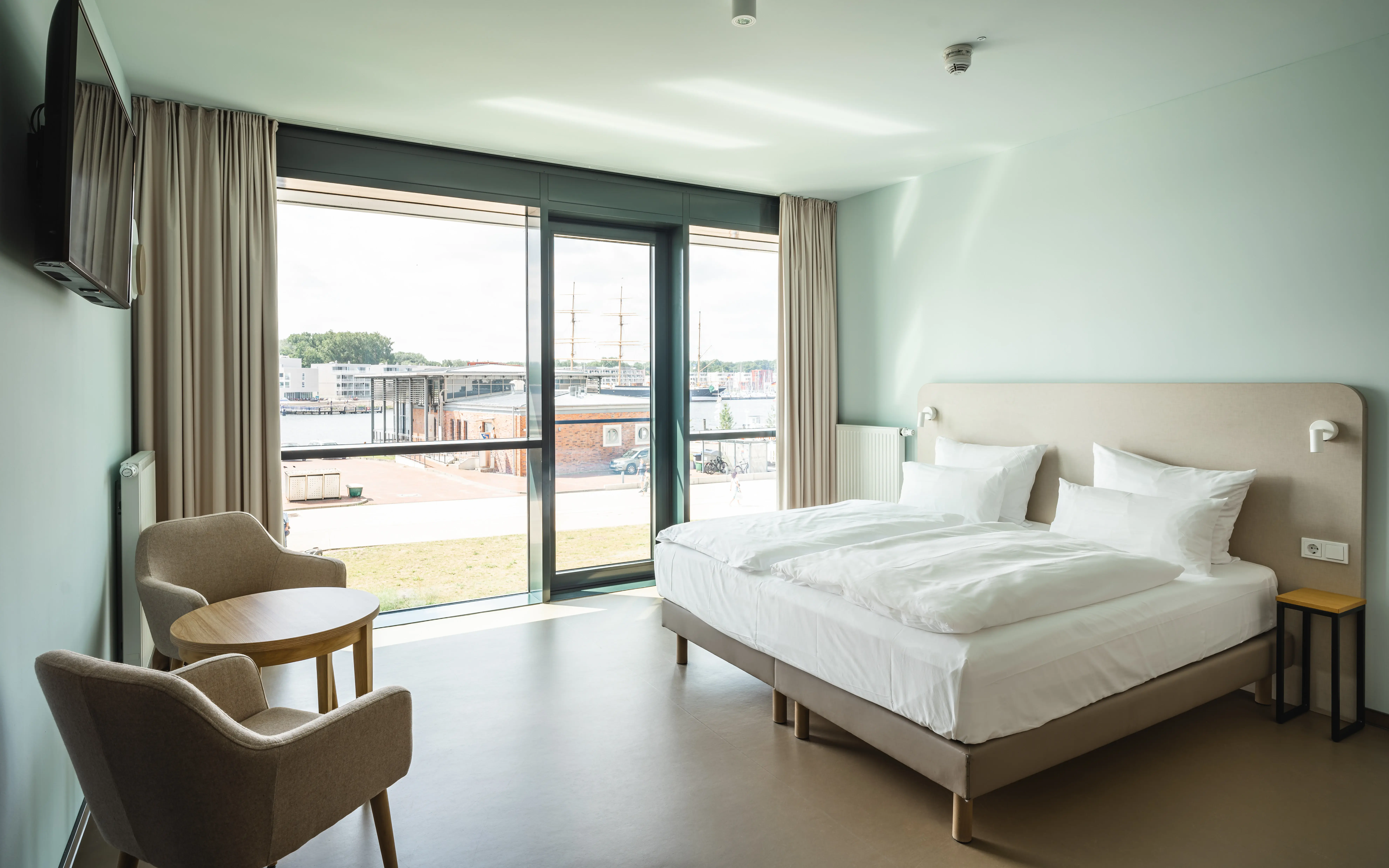 A hotel room with minimalistic decor, featuring a double bed, light furniture, and a large window overlooking a harbor with docked ships.