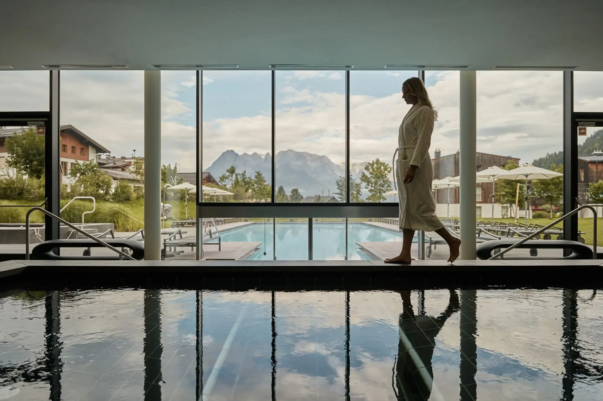 Woman walks past an indoor pool and in the background you can see the outdoor pool and a beautiful mountain backdrop.