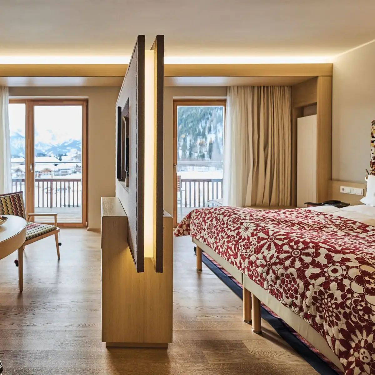 Modernly furnished hotel room with separate living and sleeping areas, wooden flooring and warm colours. A sofa with patterned cushions, a round wooden table with two glasses of wine and a TV on a partition wall dominate the room.