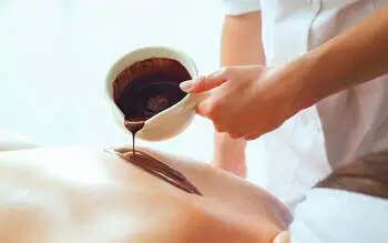 A person pours chocolate onto the back of another person in an indoor room.