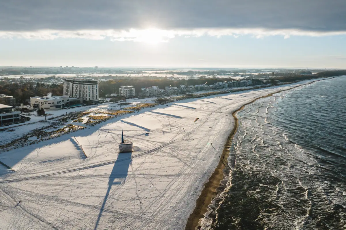 A snow-covered beach with buildings and water in winter.